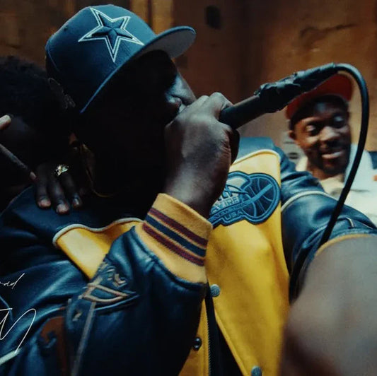 Close-up of a man in a blue and yellow vintage varsity jacket and baseball cap rapping into a microphone.